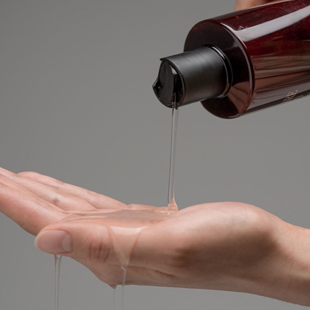 Mano che versa lo shampoo da un flacone rosso con tappo nero nel palmo. Lo shampoo è trasparente.