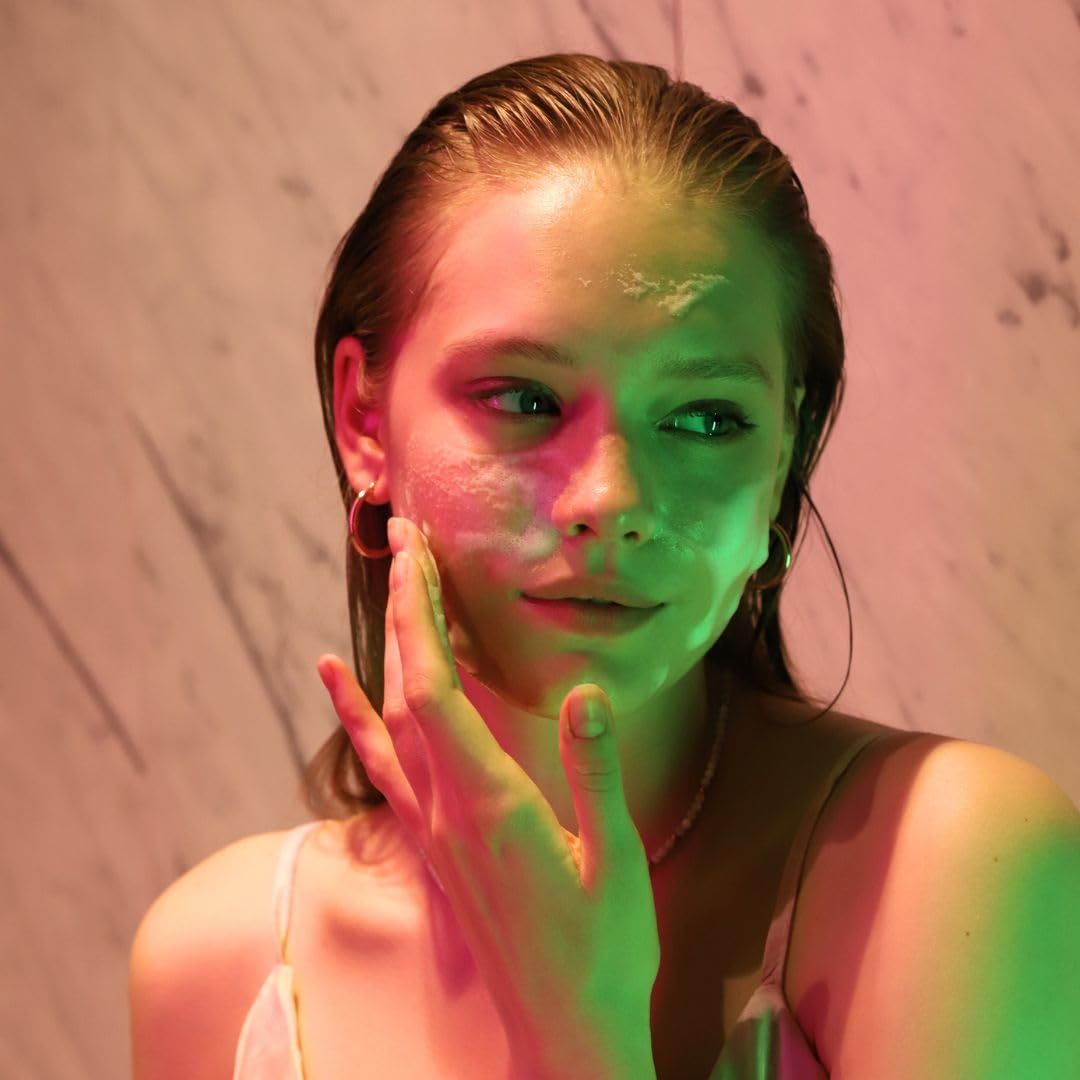 Viso di una donna con schiuma sulla pelle. Si tocca il viso. Sfondo: muro illuminato a colori.