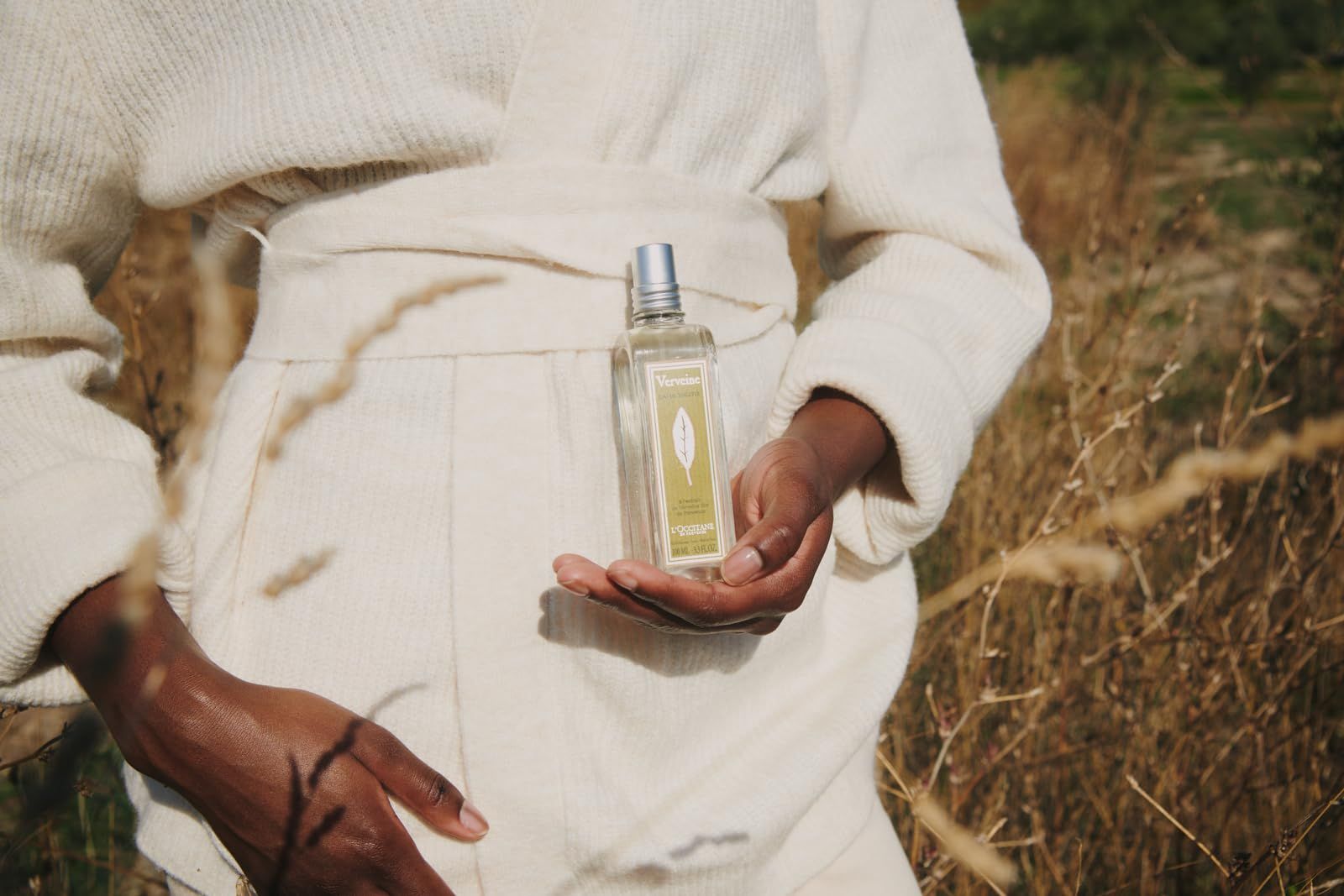 Persona che tiene in mano un flacone 'Verveine', circondata da erba. Indossa abiti chiari.