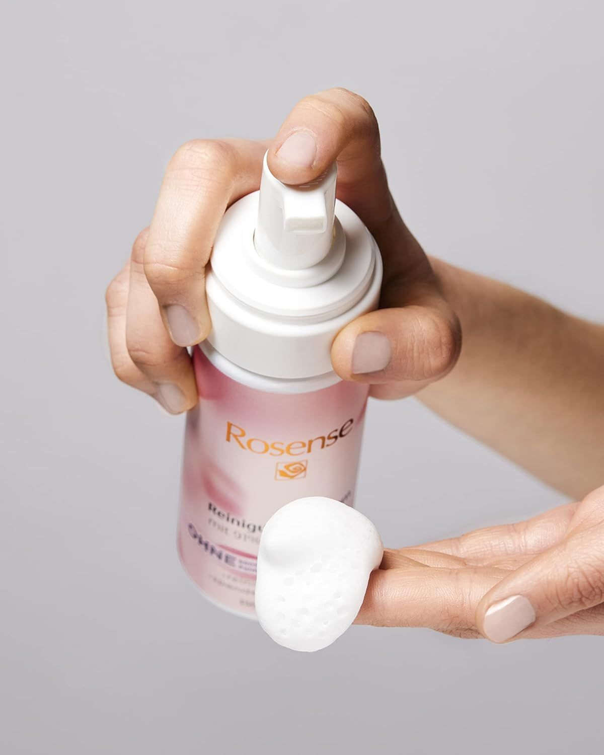 Mano che tiene un flacone di schiuma detergente Rosense e schiuma. Erogatore bianco, etichetta rosa. Schiuma sulla mano.