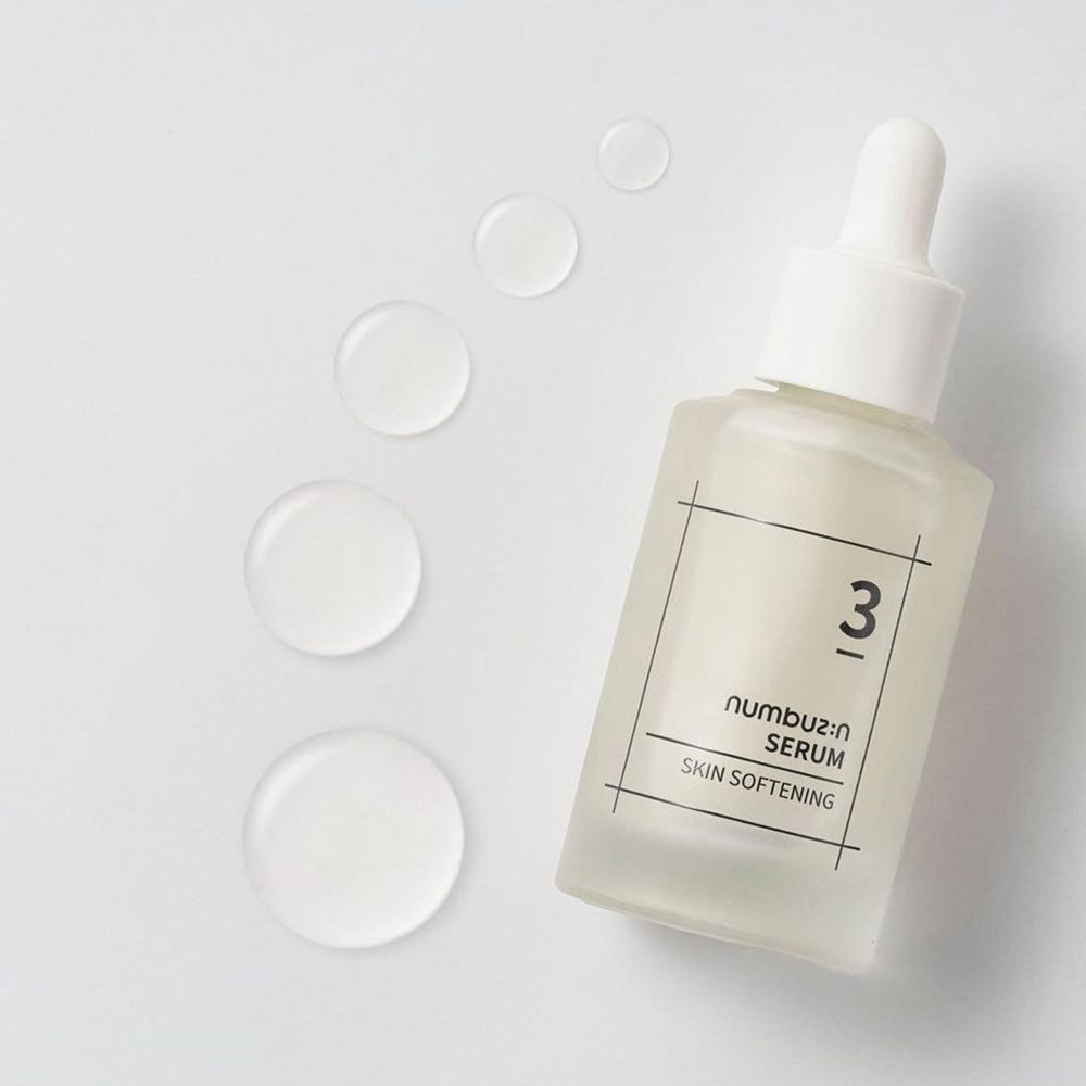 Flacone di siero con tappo e contagocce bianchi. Scritta: 3, Serum, Skin Softening. Diverse gocce di siero accanto. Flacone in vetro trasparente e opaco.