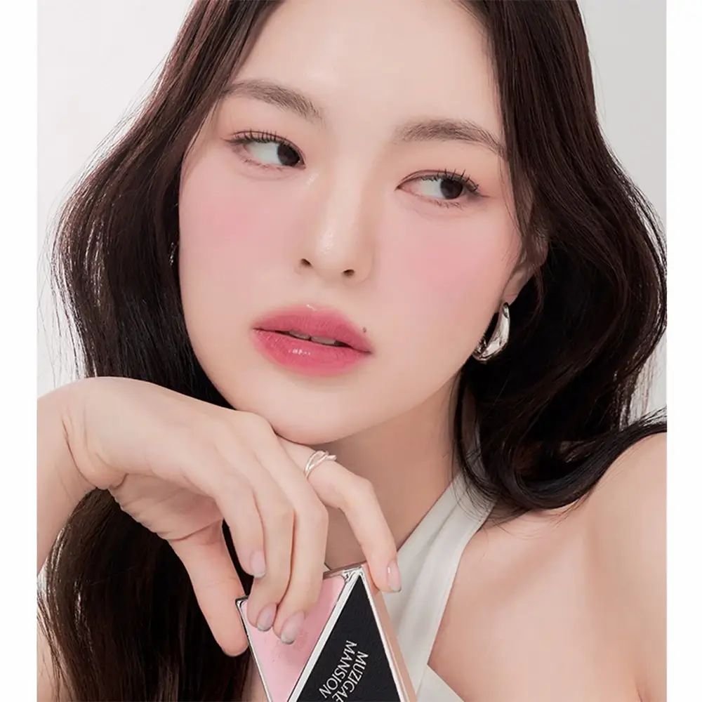 Donna con fard sulle guance. Tiene in mano una palette quadrata MUZIGAE MANSION Fitting Blush. Fard in polvere rosa in un astuccio nero e argento.