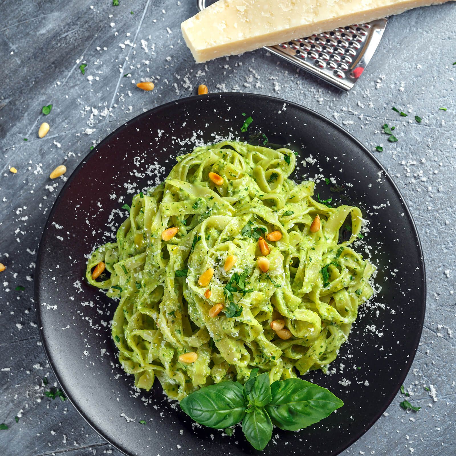 Piatto nero con tagliatelle verdi, pesto, pinoli e parmigiano. Su sfondo grigio.