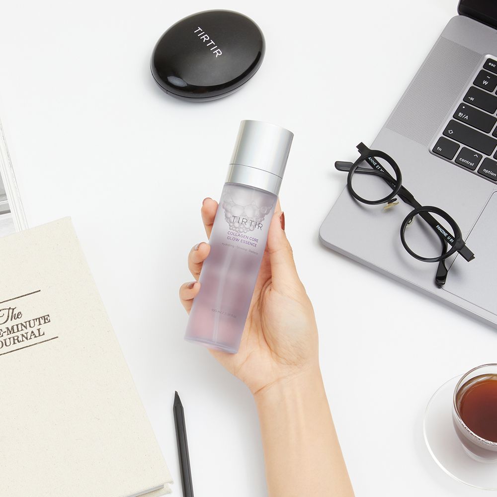 Una mano tiene TIRTIR Collagen Core Glow Essence accanto a un taccuino e occhiali.