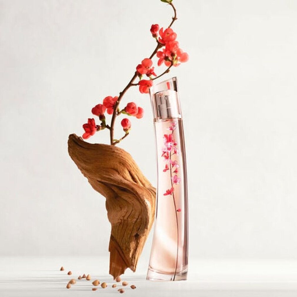Flacone di profumo con design floreale, accanto a legno e ramo fiorito. Flacone con contenuto rosa e motivo floreale.