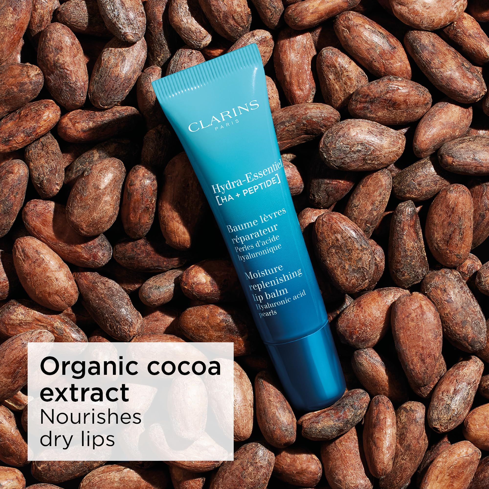 Tubo di balsamo labbra Clarins Hydra-Essentiel su fave di cacao. Testo: Hydra-Essentiel, Baume lèvres réparateur, Moisture replenishing lip balm.