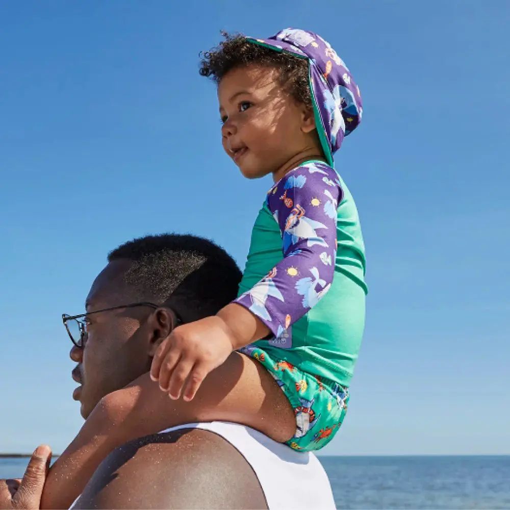 Bambino sulle spalle di una persona, indossa un costume da bagno. Costume verde-viola e cappello da sole. In spiaggia.