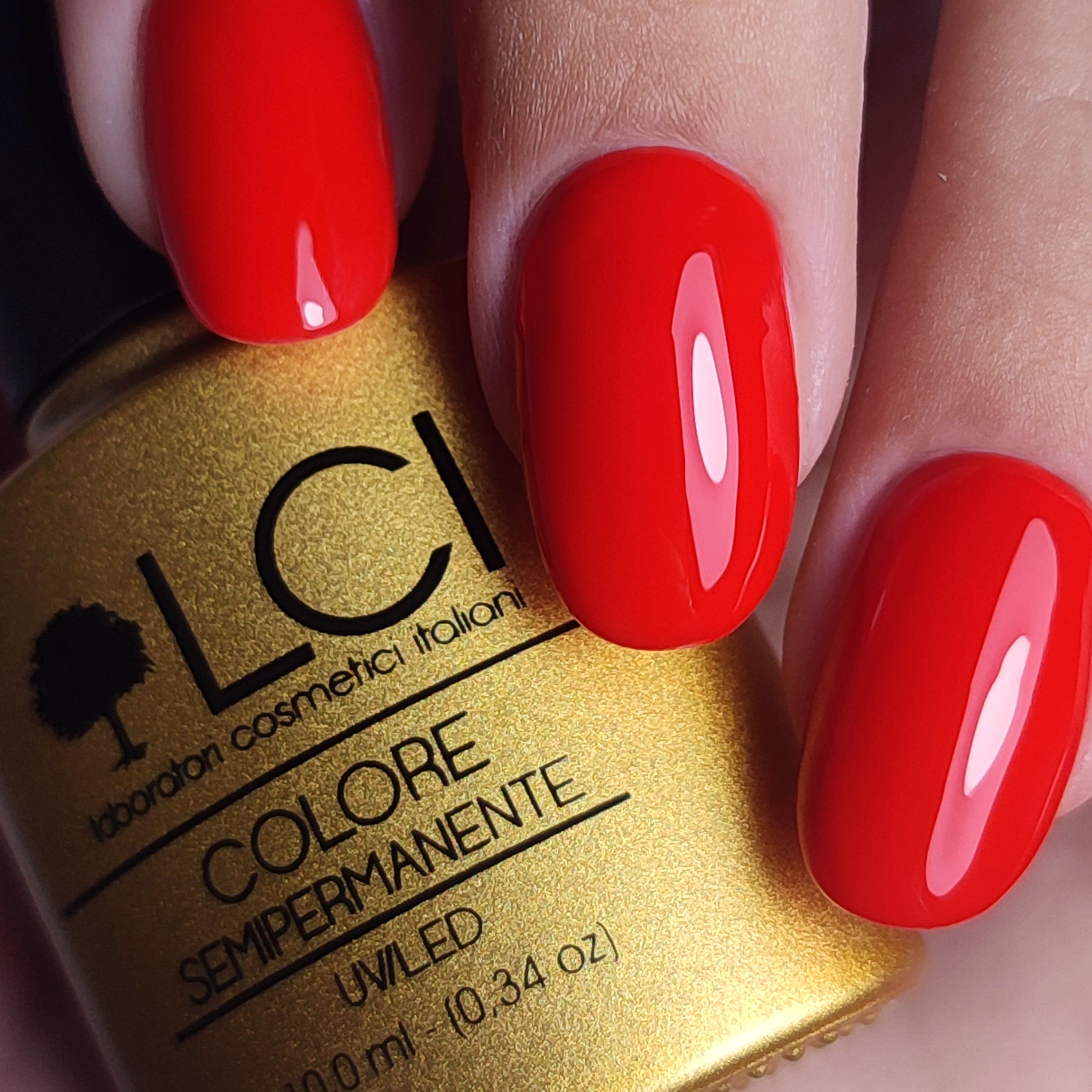 Primo piano di unghie rosse smaltate accanto a un flacone di smalto dorato. Scritta: LCI, COLORE SEMIPERMANENTE.