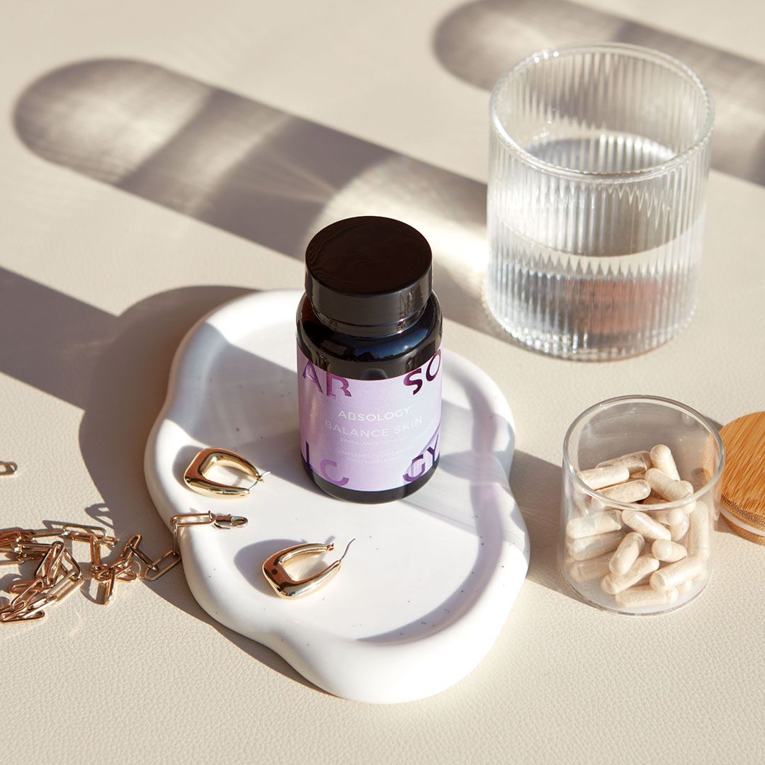 Flacone Absology Balance Skin su piatto bianco. Bicchiere d'acqua, capsule in vetro. Orecchini dorati.