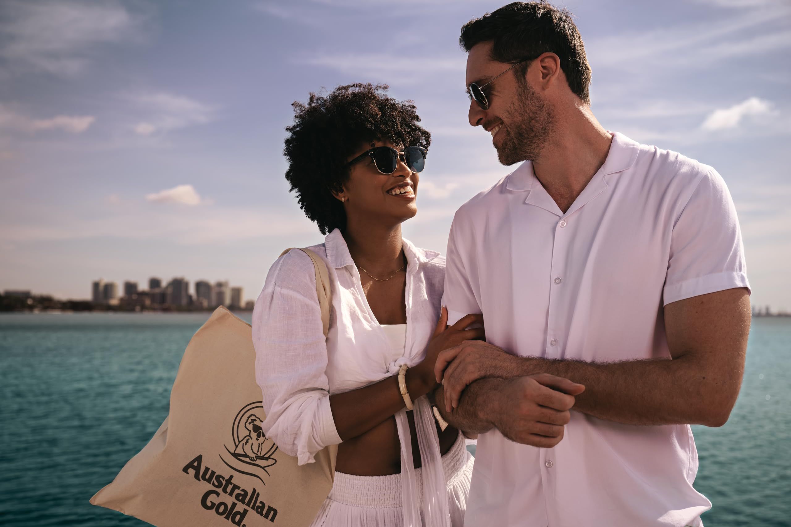 Coppia al mare. Donna con occhiali da sole e uomo con occhiali da sole. Entrambi in bianco. Borsa con logo.