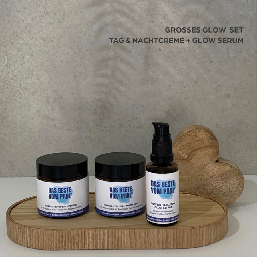 Tre prodotti su un vassoio. Scritta: Das Beste vom Paul. Grande set glow. Crema giorno e notte, siero. Cuore di legno come decorazione.