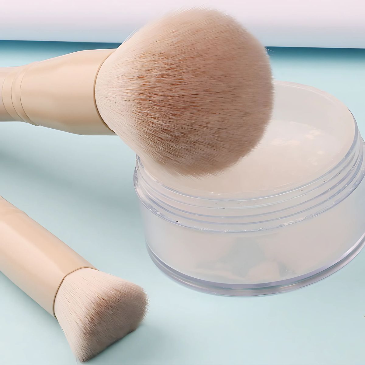 Due pennelli per il trucco con manici beige chiaro accanto a un contenitore trasparente con polvere bianca.
