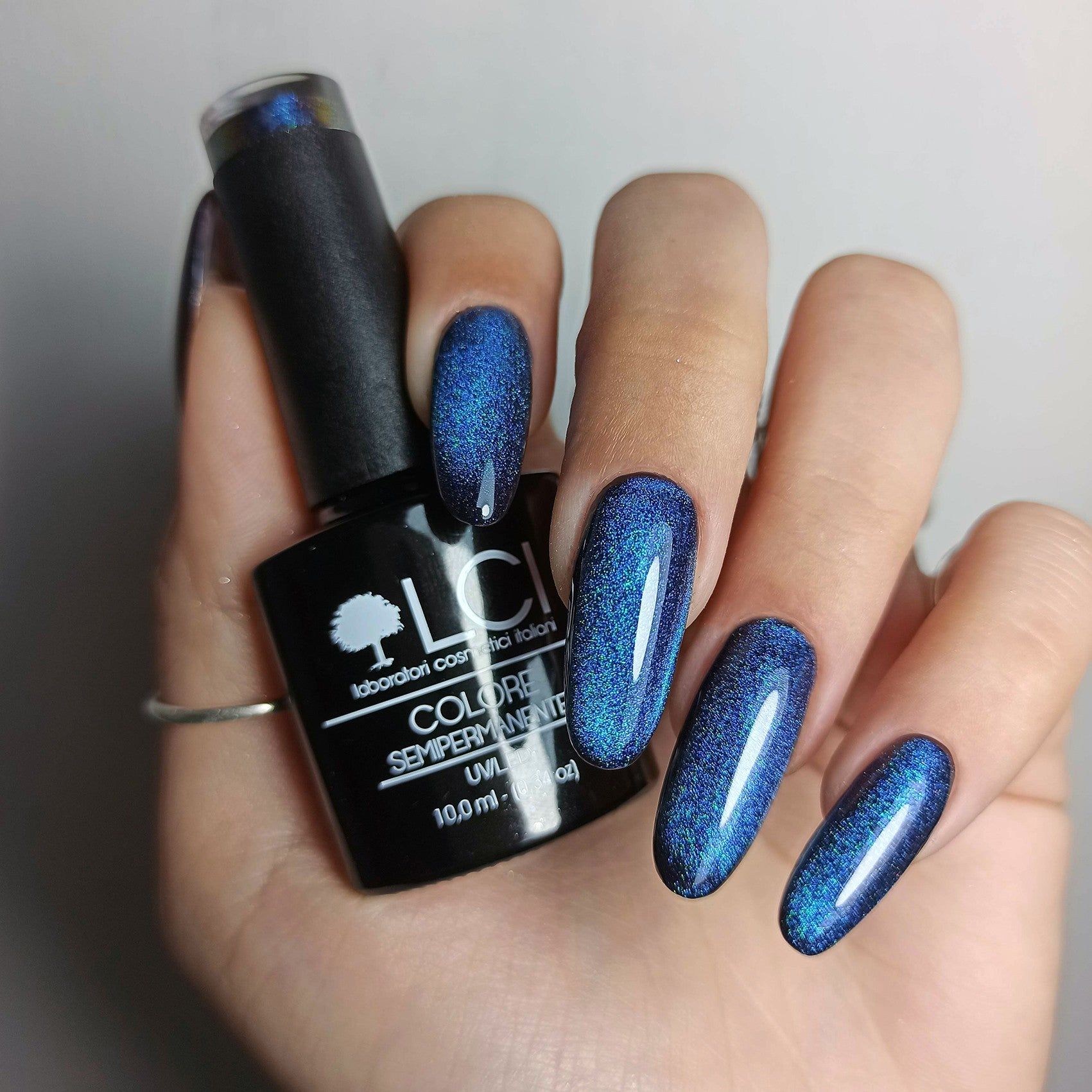 Mano con unghie smaltate di blu. Flacone di smalto con scritta: LCI, COLORE SEMIPERMANENTE, UVLED 10,0 ml.