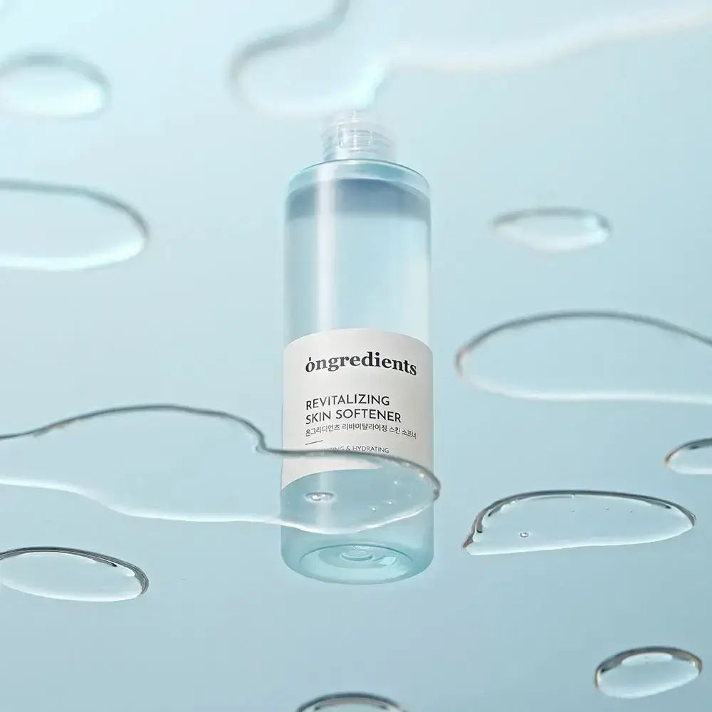 Contenitore cilindrico trasparente con etichetta bianca. Scritta: òngredients, Revitalizing Skin Softener. Circondato da gocce d'acqua.