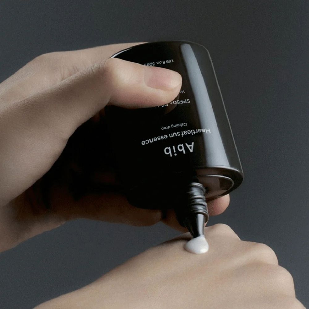 Mani che spremono crema da un flacone nero. Scritta: Abib, Heartleaf sun essence, SPF50+ PA++++.