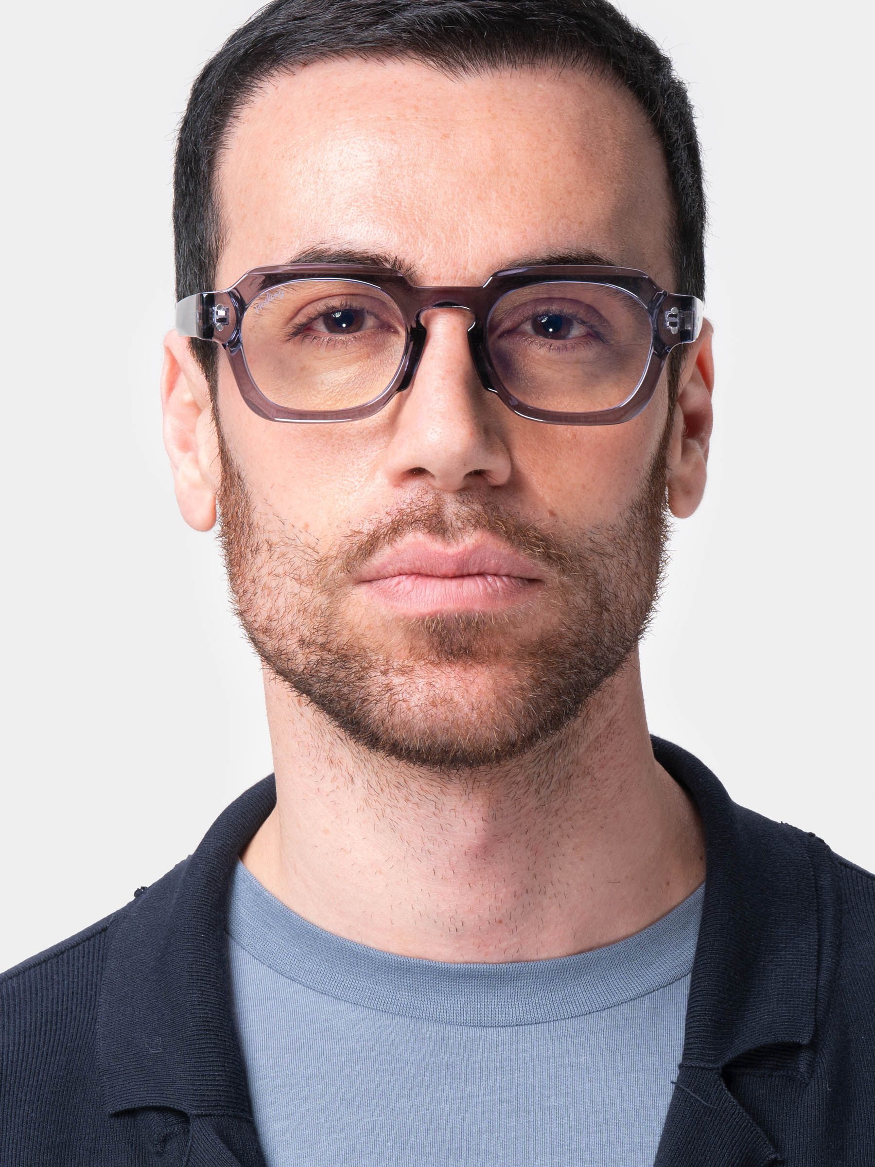 Uomo con occhiali. Montatura grigio-viola, lenti rettangolari. Ponte nasale nero. Logo Herling sull'asta. Camicia blu scuro.