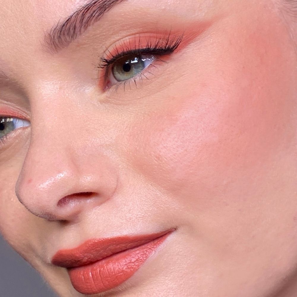 Primo piano di un viso con occhi, guance e labbra truccate. Rossetto color corallo. Tono della pelle chiaro.