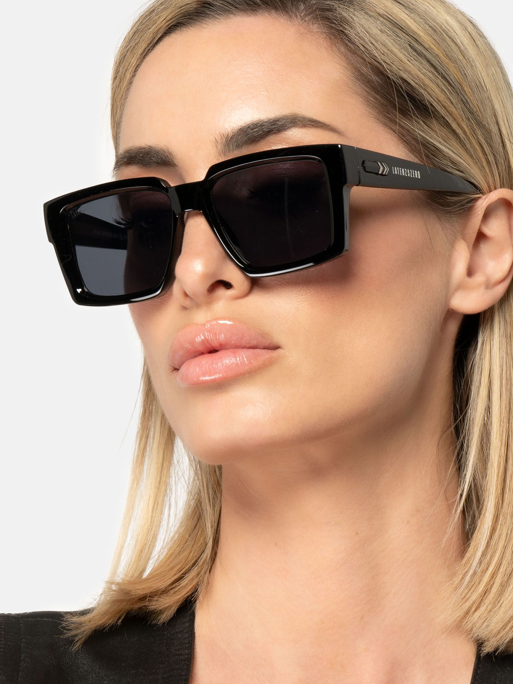 Donna con occhiali da sole neri. Lenti rettangolari, montatura ampia. Logo Latenzazero sull'asta.