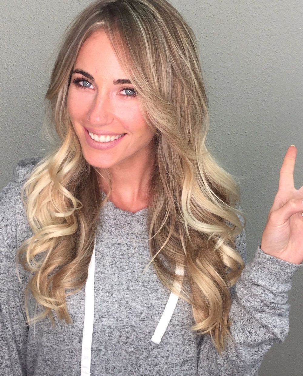 Ritratto di una donna sorridente con capelli biondi. Mostra il segno della vittoria.