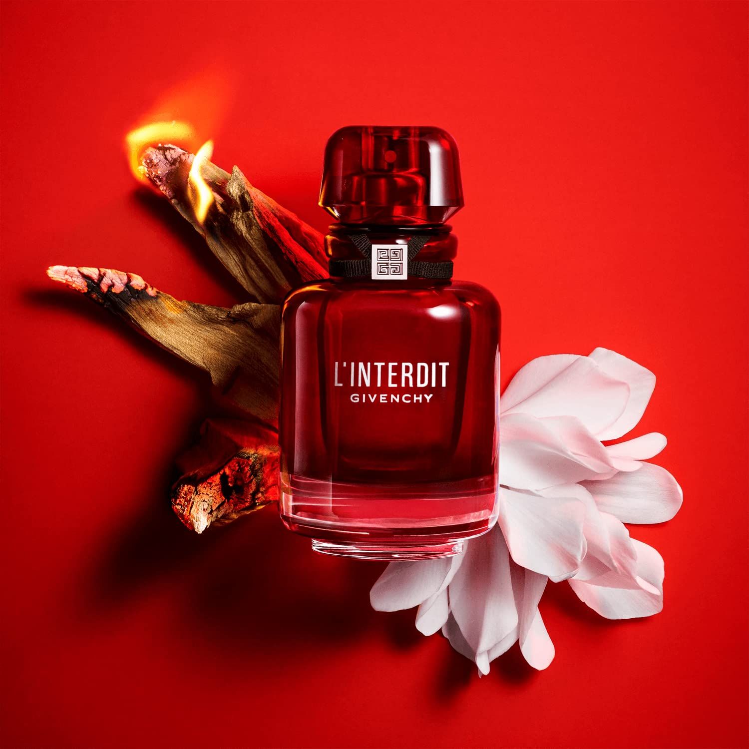 Flacone di profumo rosso con nastro nero e tappo argentato, circondato da fiamme, legno e un fiore bianco.