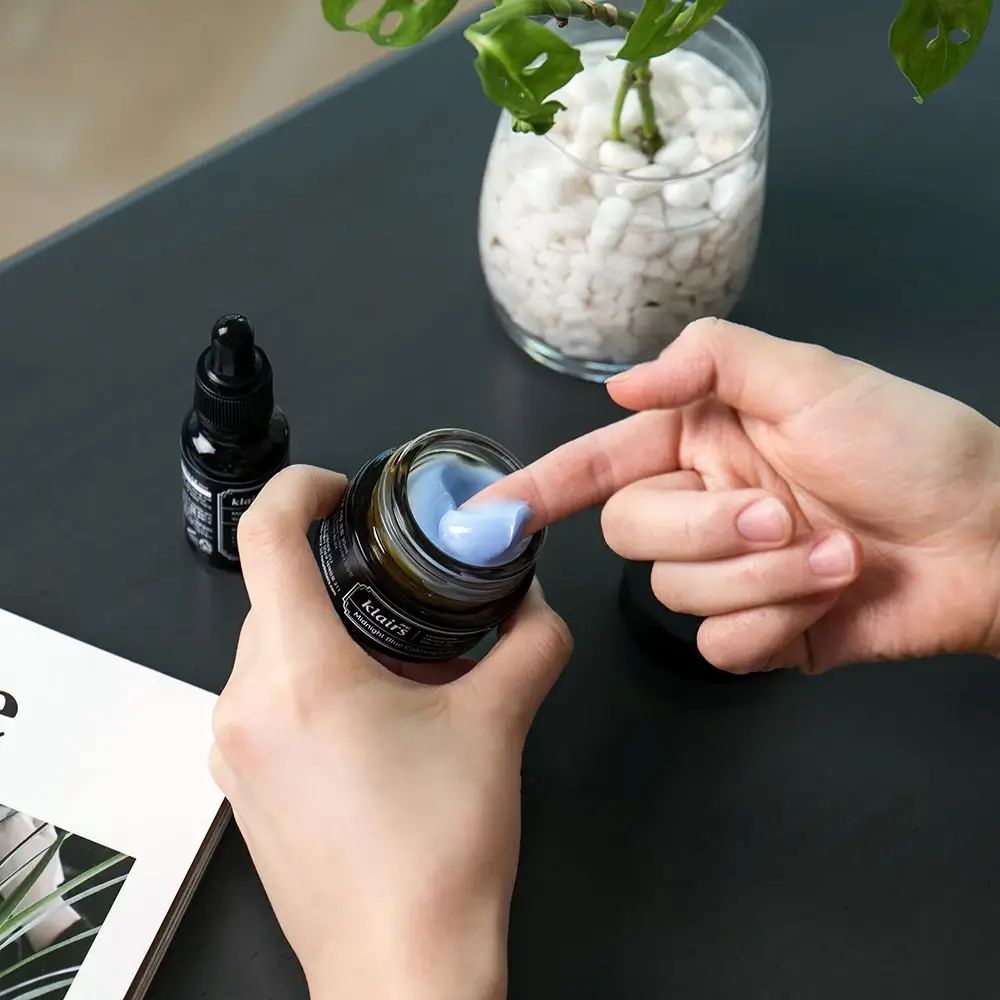 Mani che tengono un vasetto aperto di crema blu. Accanto, una bottiglia con etichetta nera.