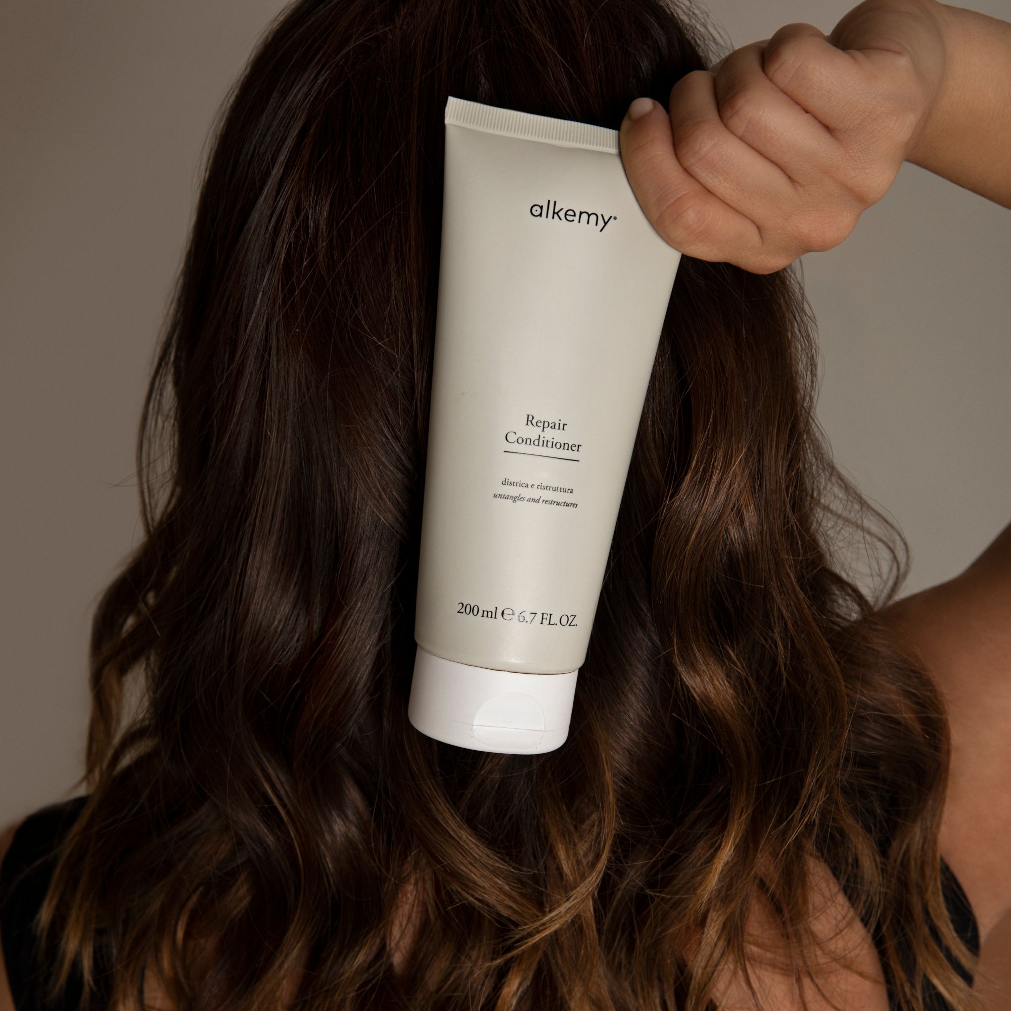 Un tubetto di Balsamo Riparatore Alkemy, 200 ml, tenuto dietro i capelli di una persona.
