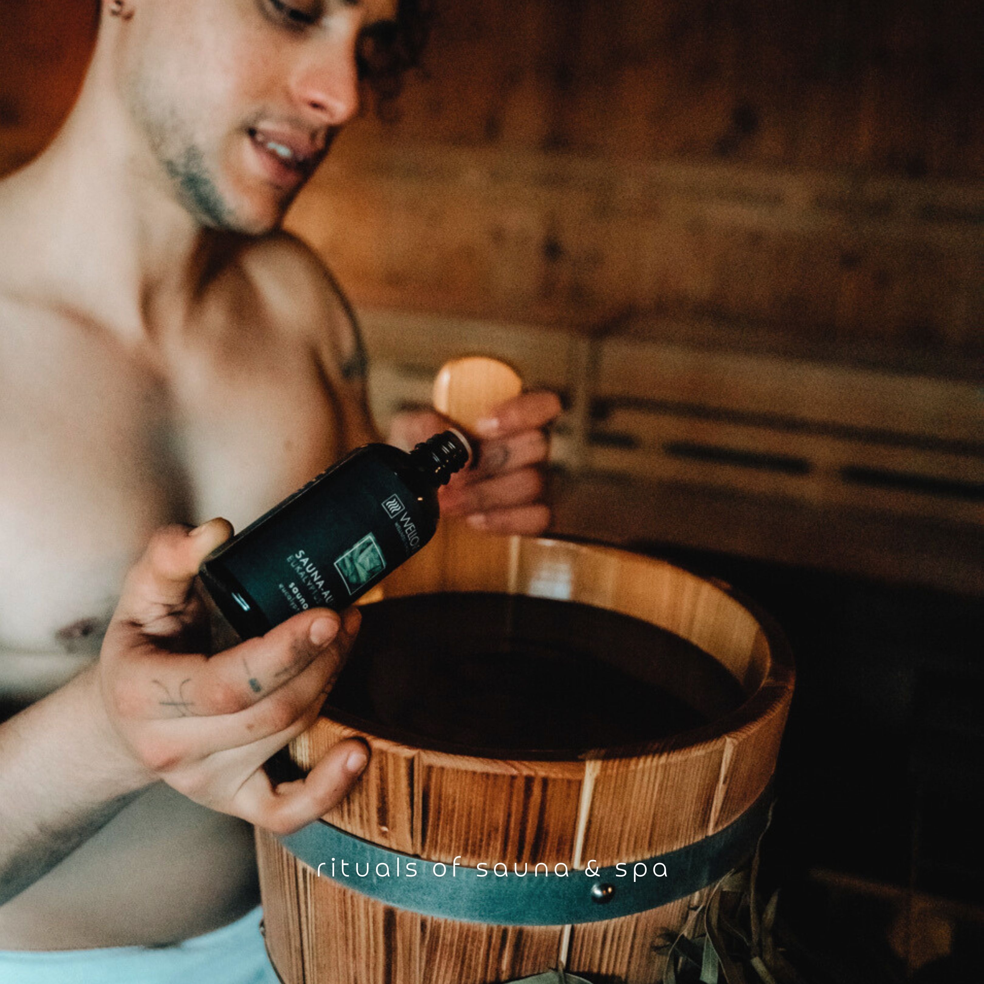 Uomo in sauna, tiene in mano una bottiglia e un secchio di legno. Scritta: Sauna-Aufguss.