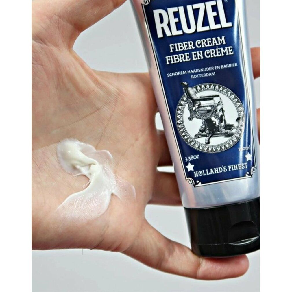 Mano con crema. Tubo con etichetta blu. Scritta REUZEL FIBER CREAM. Immagine di una poltrona da barbiere.