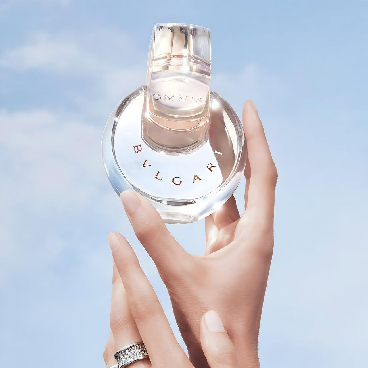 Flacone di profumo tra le mani di una donna davanti a un cielo blu. Il flacone è rotondo e trasparente con accenti rosa.