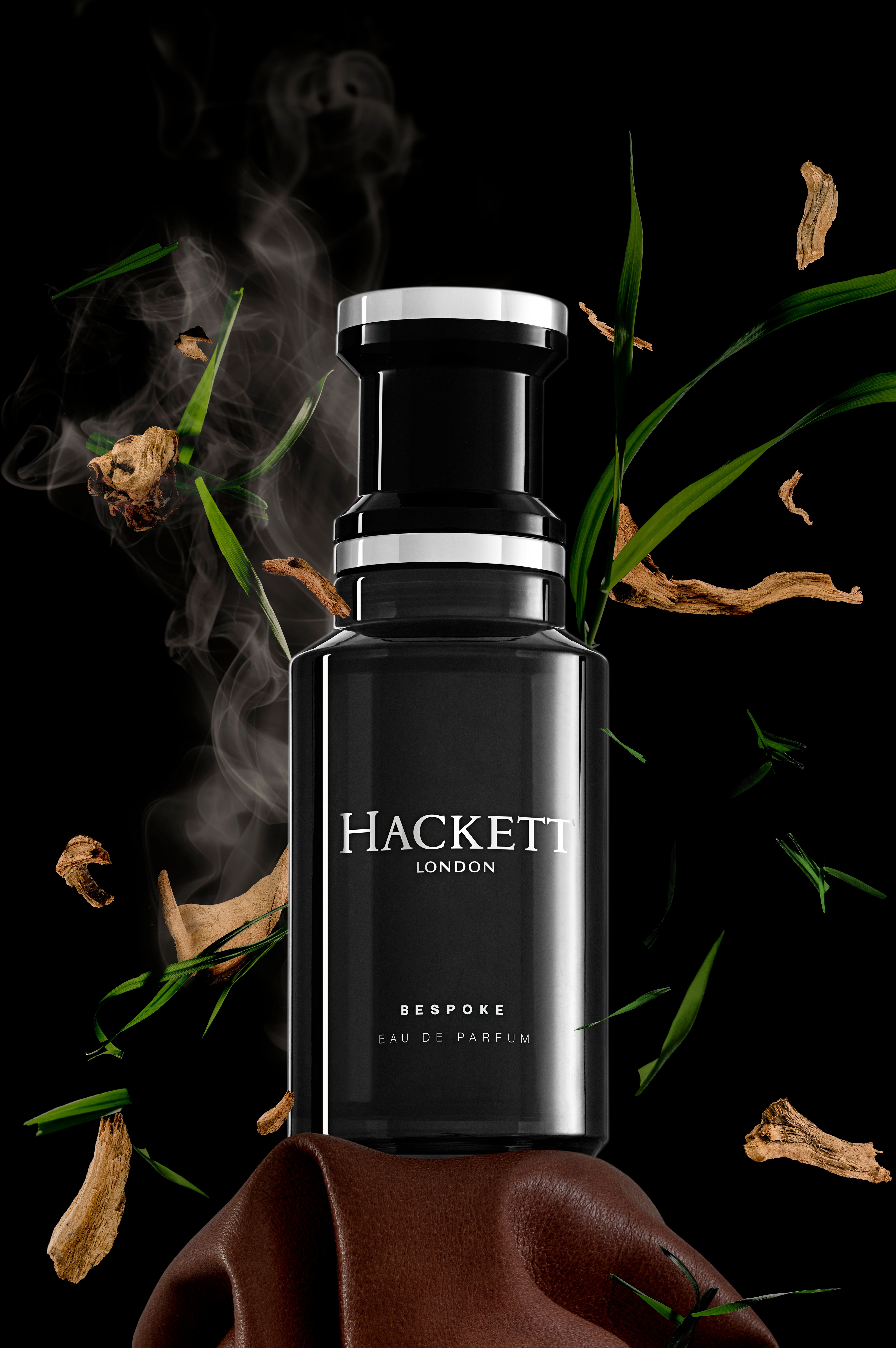 Flacone nero con tappo argentato. Hackett Bespoke Eau de Parfum. Ambiente: piante, fumo. Sfondo nero.