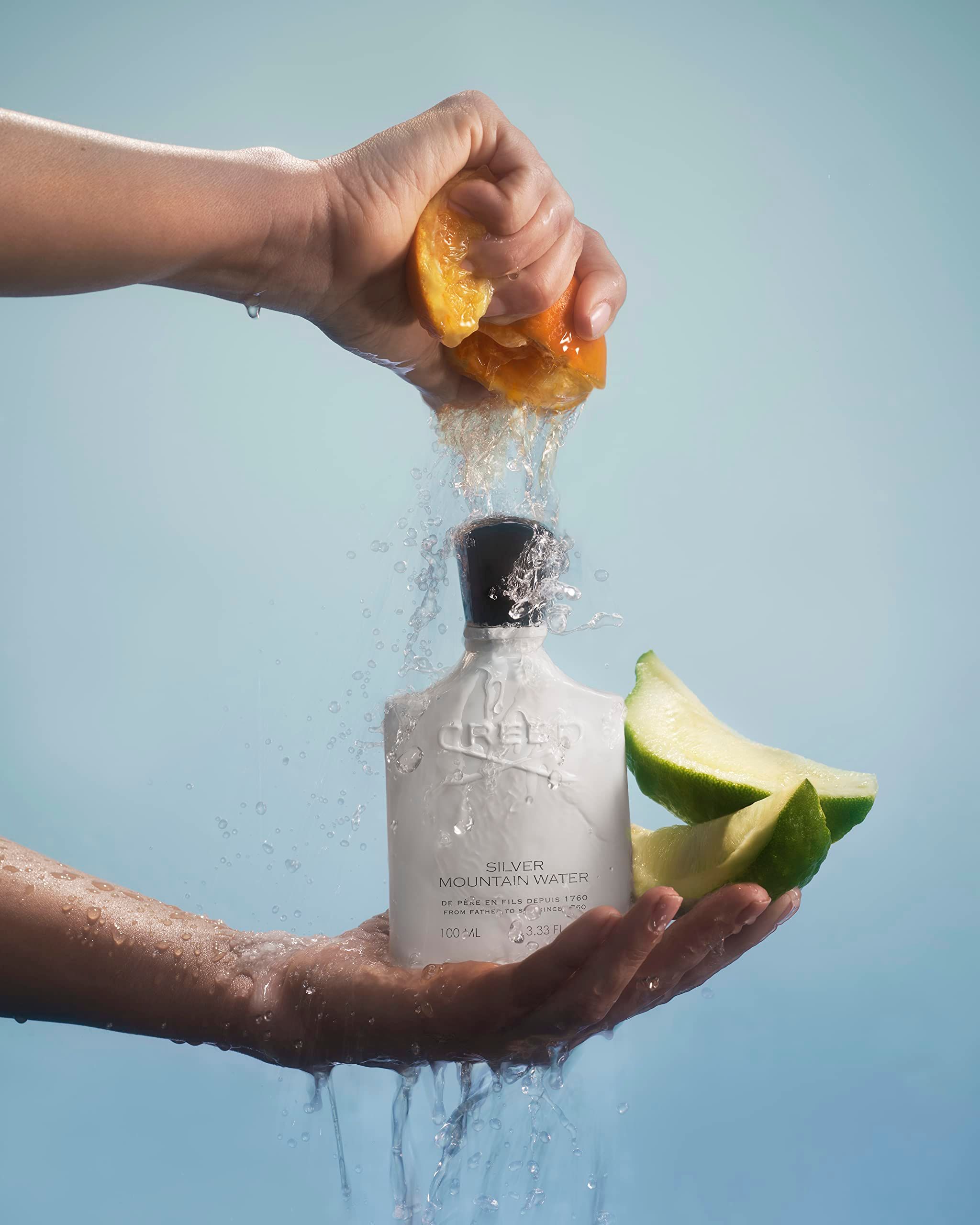 Mani che spremono un'arancia sopra un flacone di profumo. Scritta: Creed Silver Mountain Water.