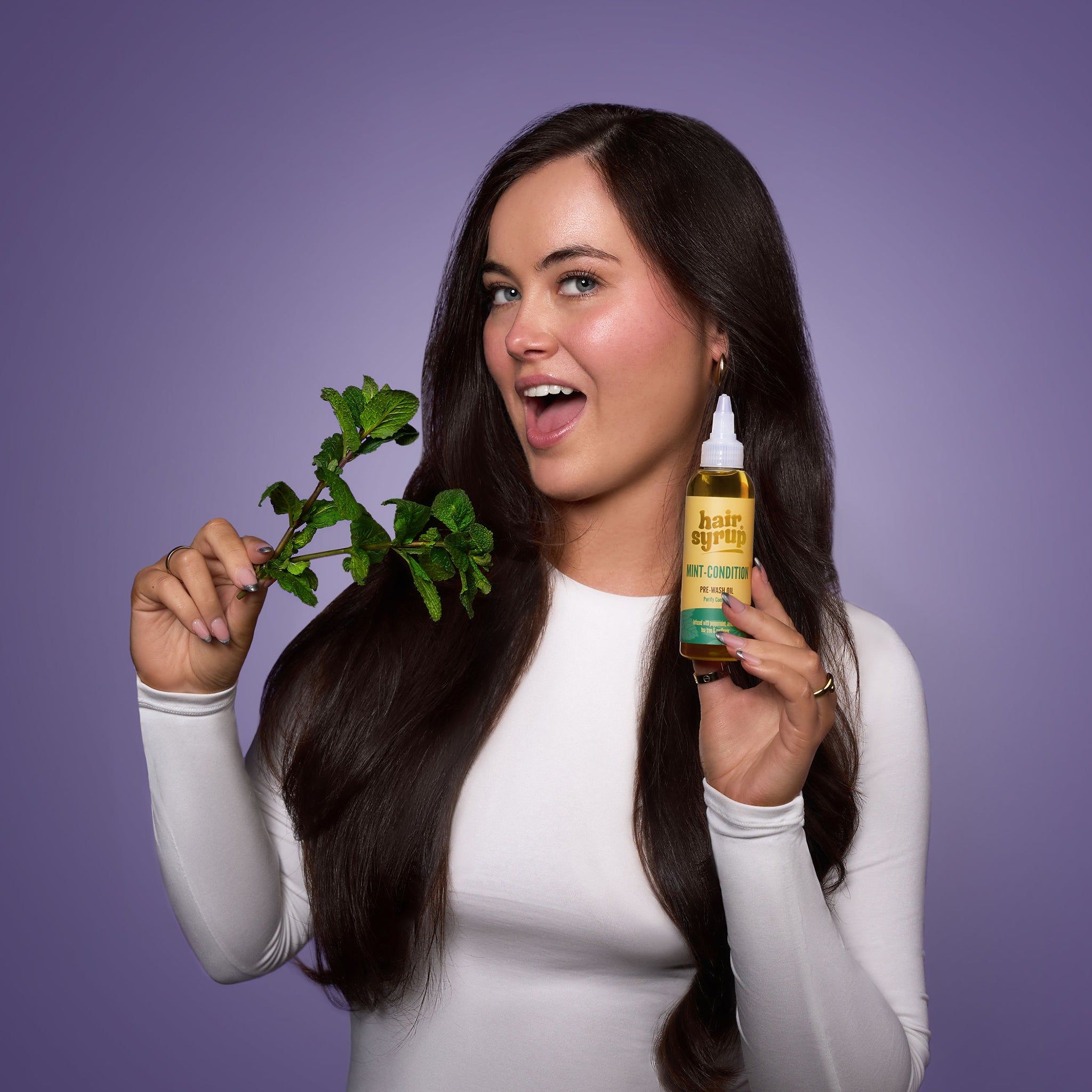 Donna con capelli lunghi tiene flacone "Hair Syrup Mint Condition" e rametto di menta. Sfondo viola.