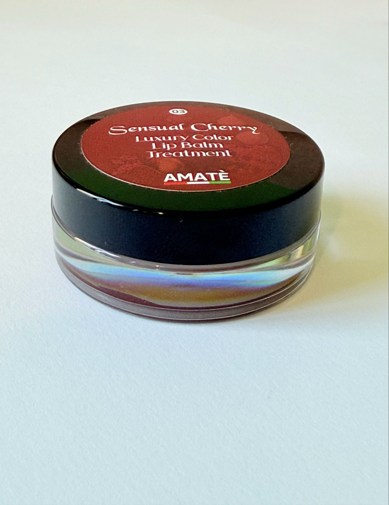 Vasetto rotondo con coperchio nero. Sul coperchio, la scritta "Sensual Cherry Luxury Color Lip Balm Treatment" e "AMATÈ".