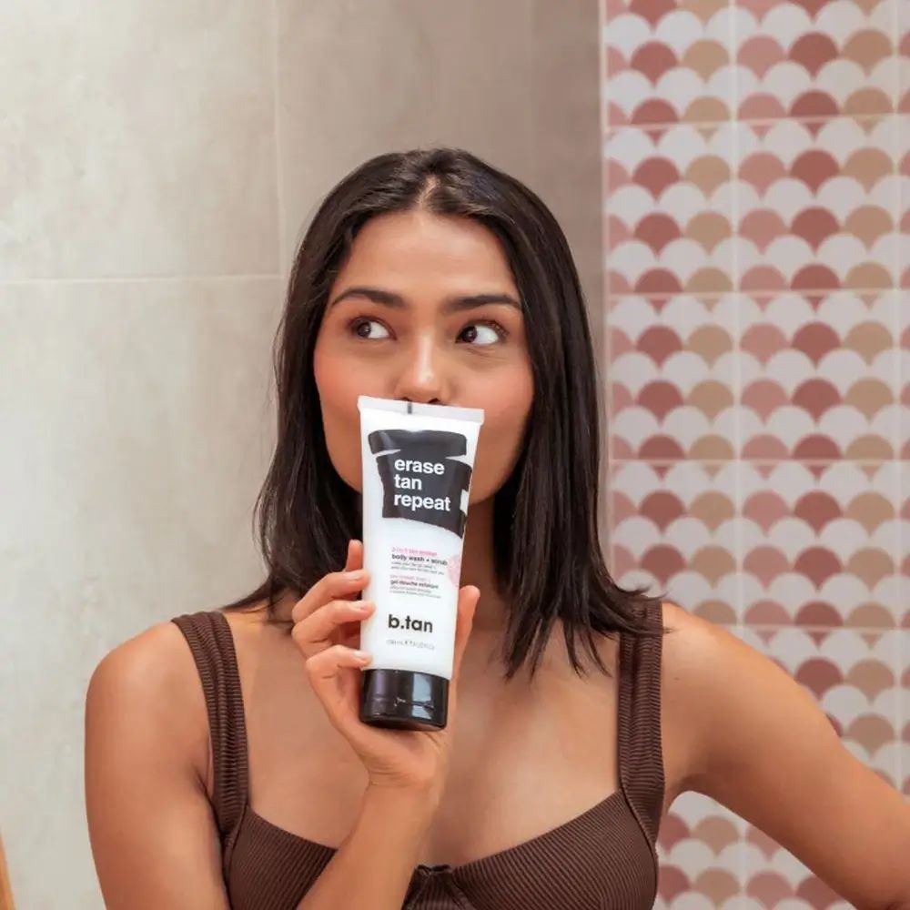 Donna tiene un tubetto davanti al viso. Sul tubetto c'è scritto "erase tan repeat" e "b.tan".