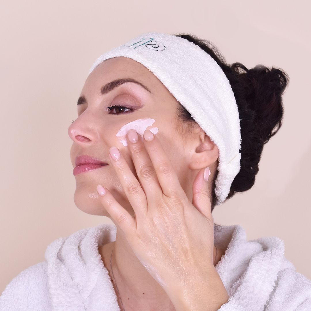 Donna applica crema sul viso. Indossa fascia e accappatoio bianchi.