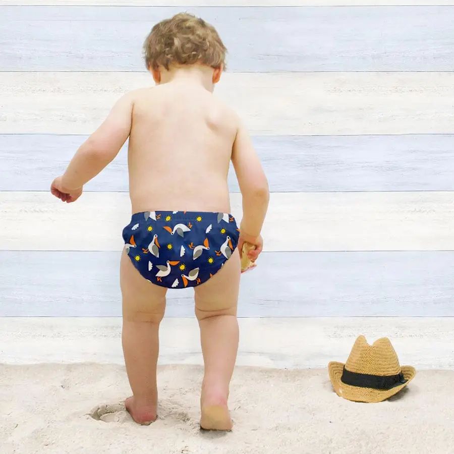 Bambino di spalle con costume contenitivo blu con stampa pellicani sulla spiaggia.
