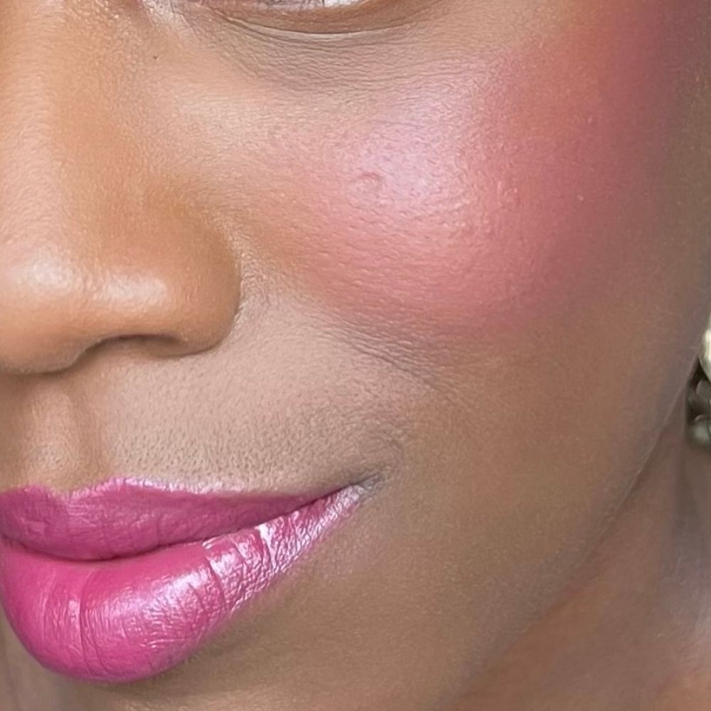 Primo piano di un viso con fard rosa sulle guance e rossetto sulle labbra. Pelle scura.