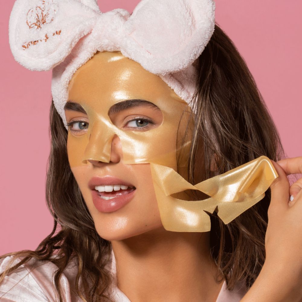 Donna con maschera viso idrogel dorata, che ne rimuove una parte. Indossa una fascia. Sfondo rosa.