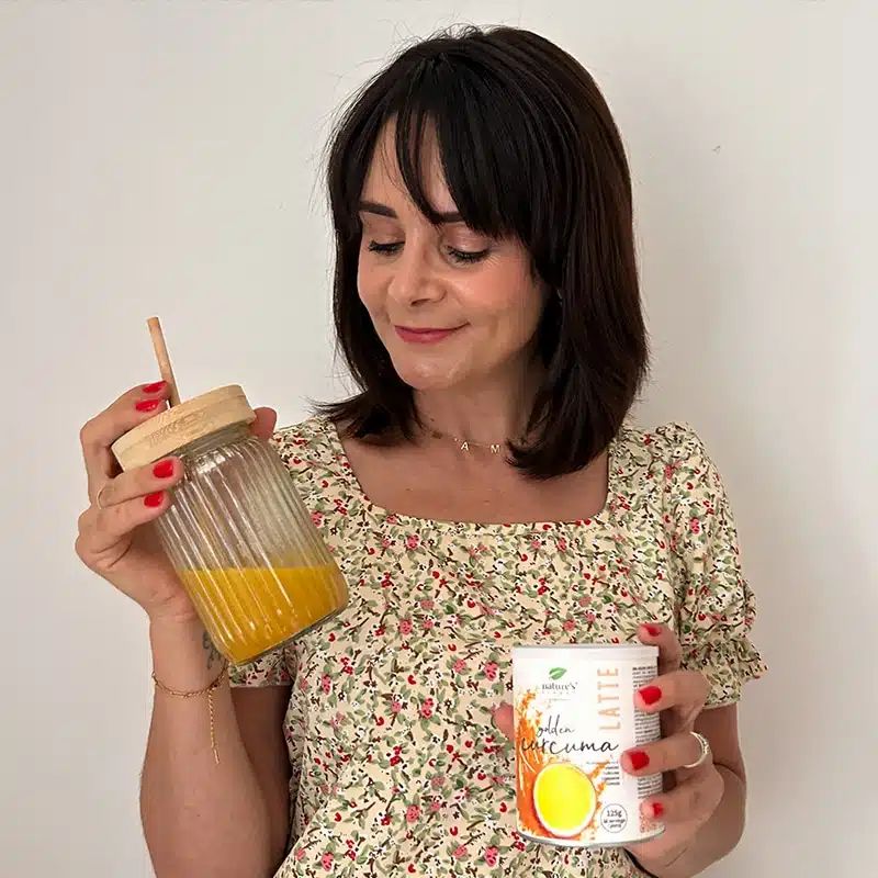 Una donna tiene un bicchiere di bevanda dorata e una lattina di Nature's Finest Golden Curcuma Latte.