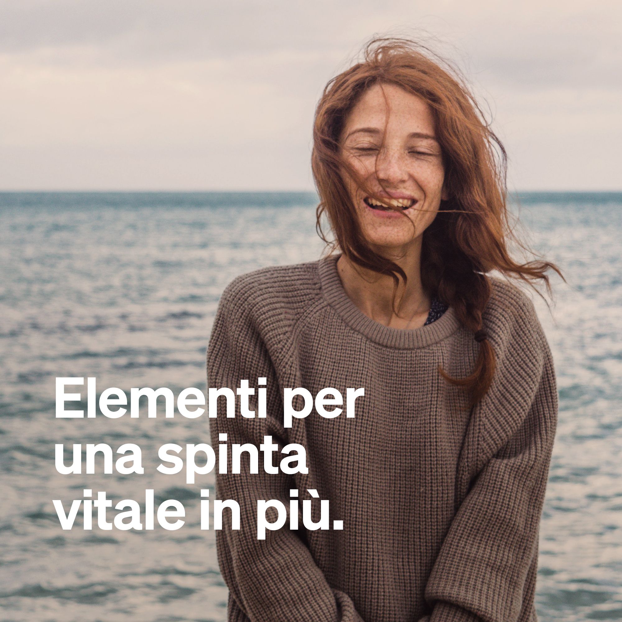 Una donna con i capelli rossi in piedi in riva al mare ride. Il testo dice "Elementi per una spinta vitale in più."