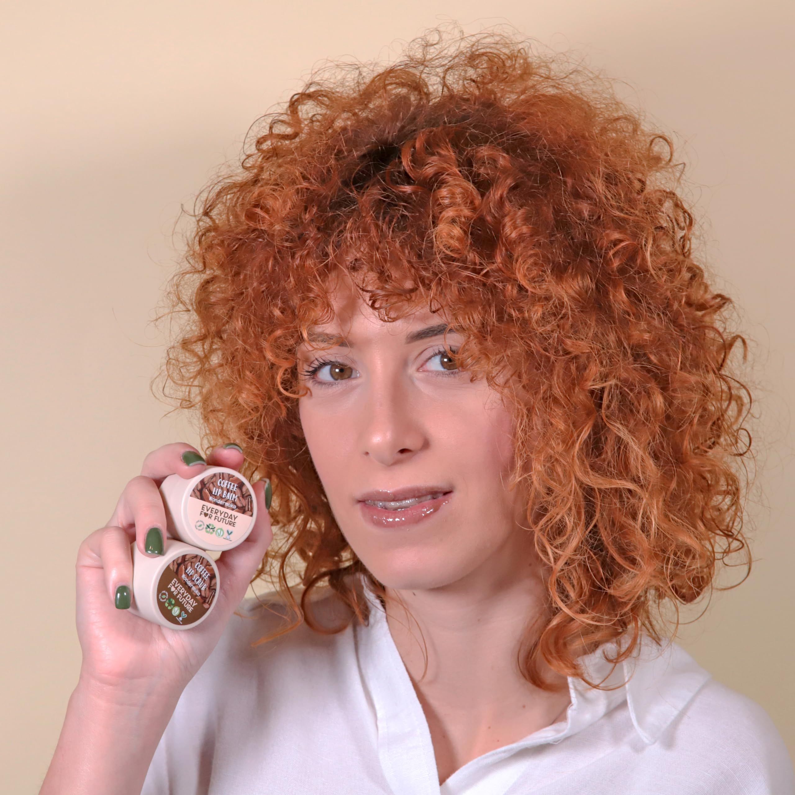 Una donna con capelli rossi ricci tiene due vasetti di scrub e balsamo labbra. Scritta: Coffee Lip Scrub & Balm.