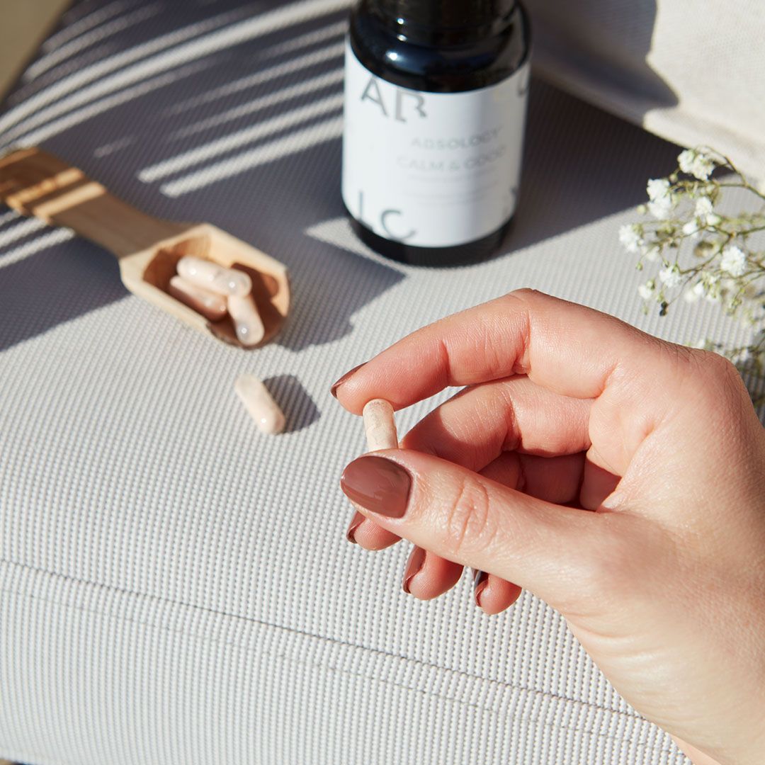 Persona tiene in mano una capsula tra le dita. Accanto, cucchiaio di legno con capsule. Su un divano.