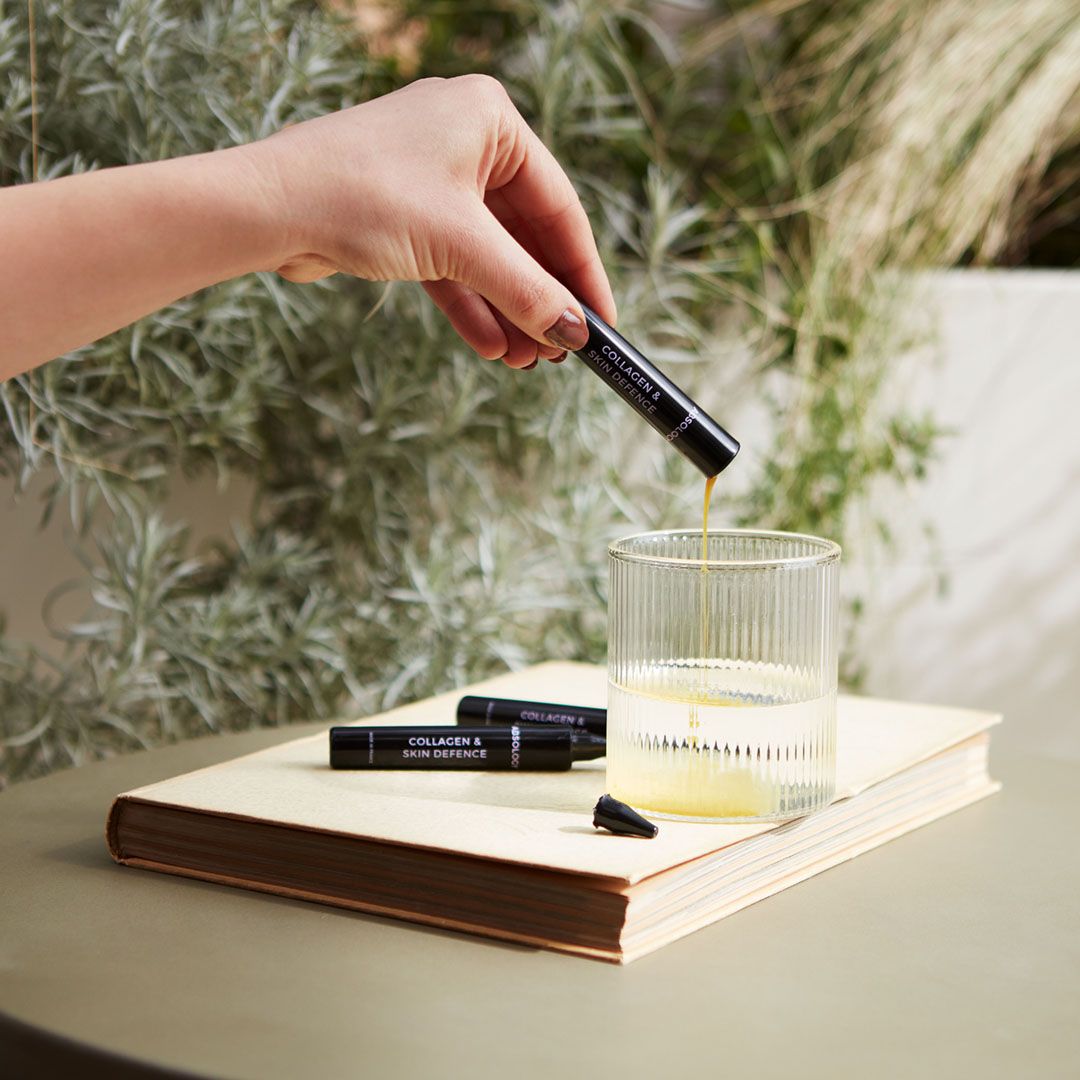 Mano versa liquido da una fiala nera in un bicchiere. Scritta: "Collagen & Skin Defence".