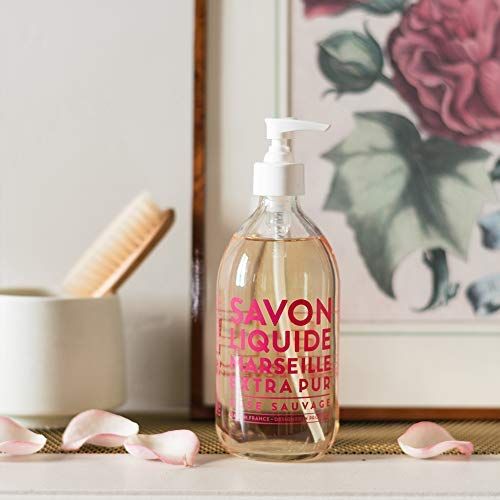 Flacone trasparente con etichetta rosa. Scritta: Savon Liquide Marseille Extra Pur Rose Sauvage. Accanto, un vaso con spazzola e petali.
