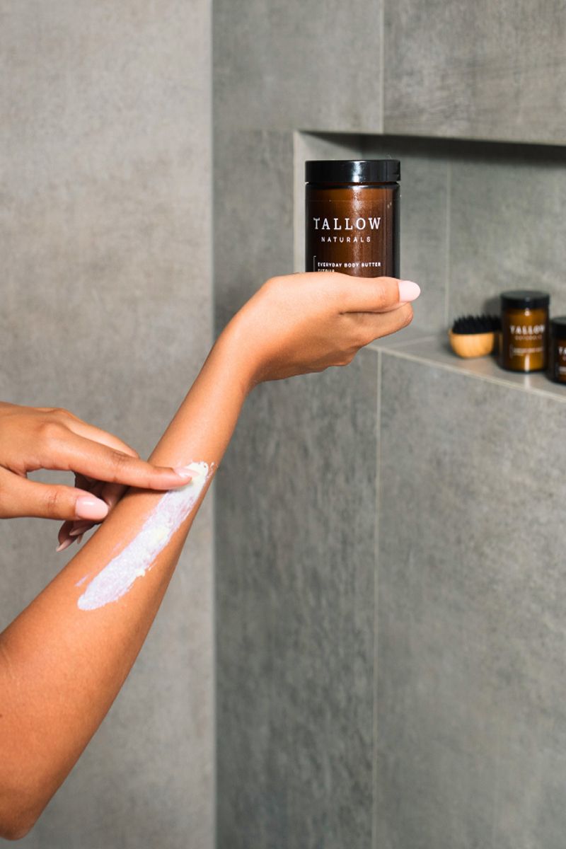 Persona applica burro corpo sul braccio. Barattolo con scritta: TALLOW NATURALS, EVERYDAY BODY BUTTER. Sfondo grigio.
