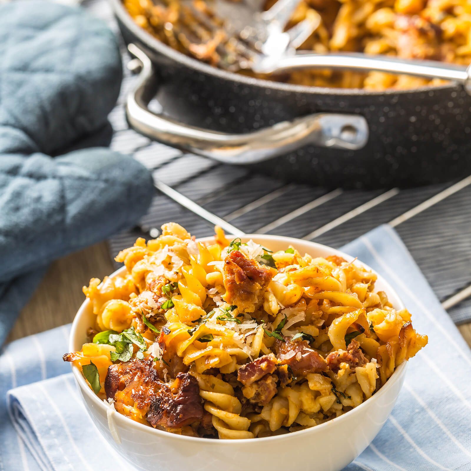 Ciotola bianca con piatto di fusilli. Contiene pomodori, pezzi di carne e formaggio. Su tessuto blu.
