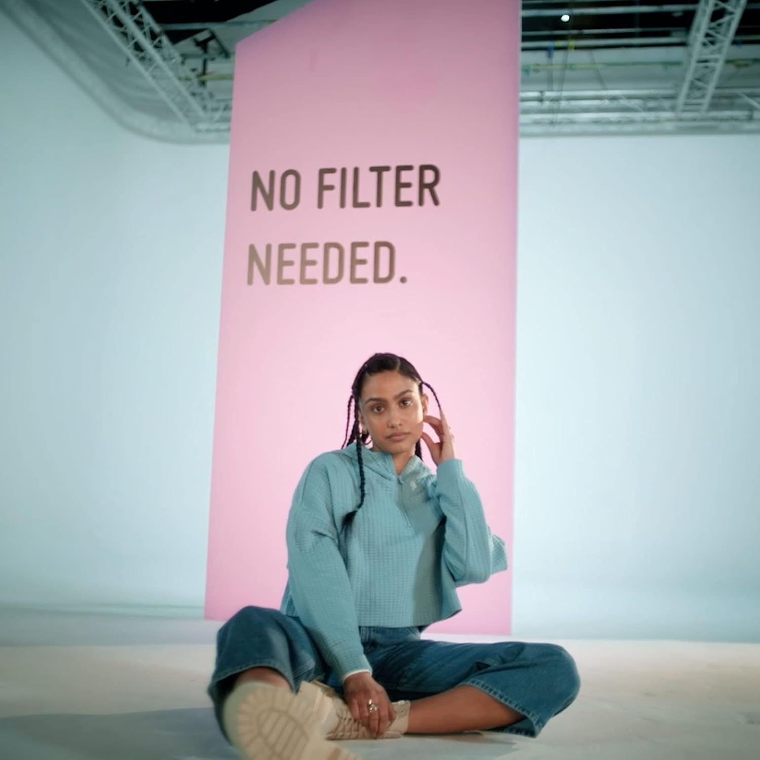 Una donna seduta davanti a uno sfondo rosa con la scritta 'NO FILTER NEEDED'. Indossa una felpa blu e jeans.
