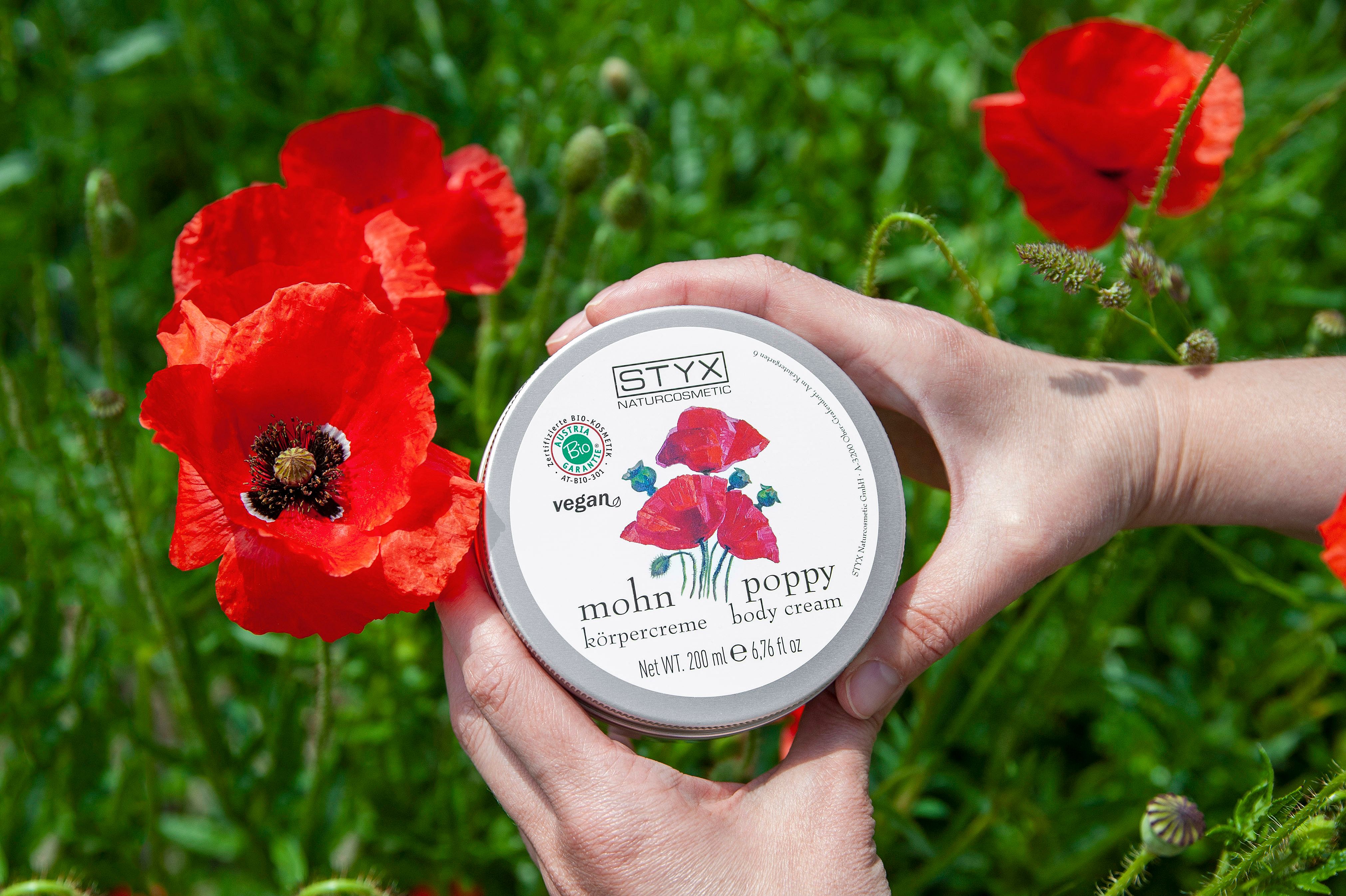 Mani che tengono un barattolo di crema con motivo papaveri. Scritta: STYX, crema corpo al papavero, vegana. Sigillo bio.