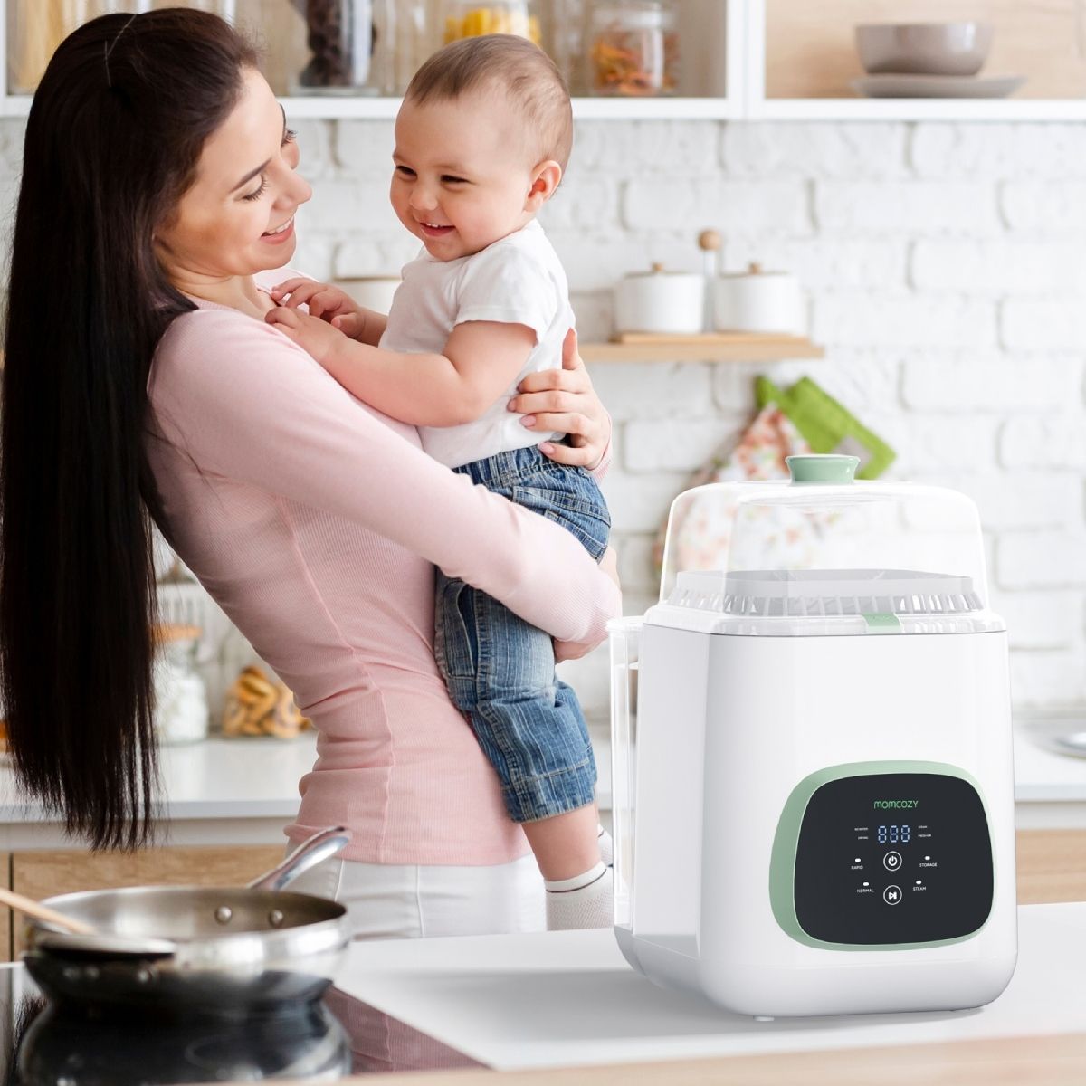 Donna tiene in braccio un bambino davanti allo sterilizzatore. Sterilizzatore bianco con dettagli verdi. Logo Momcozy visibile. Cucina sullo sfondo.