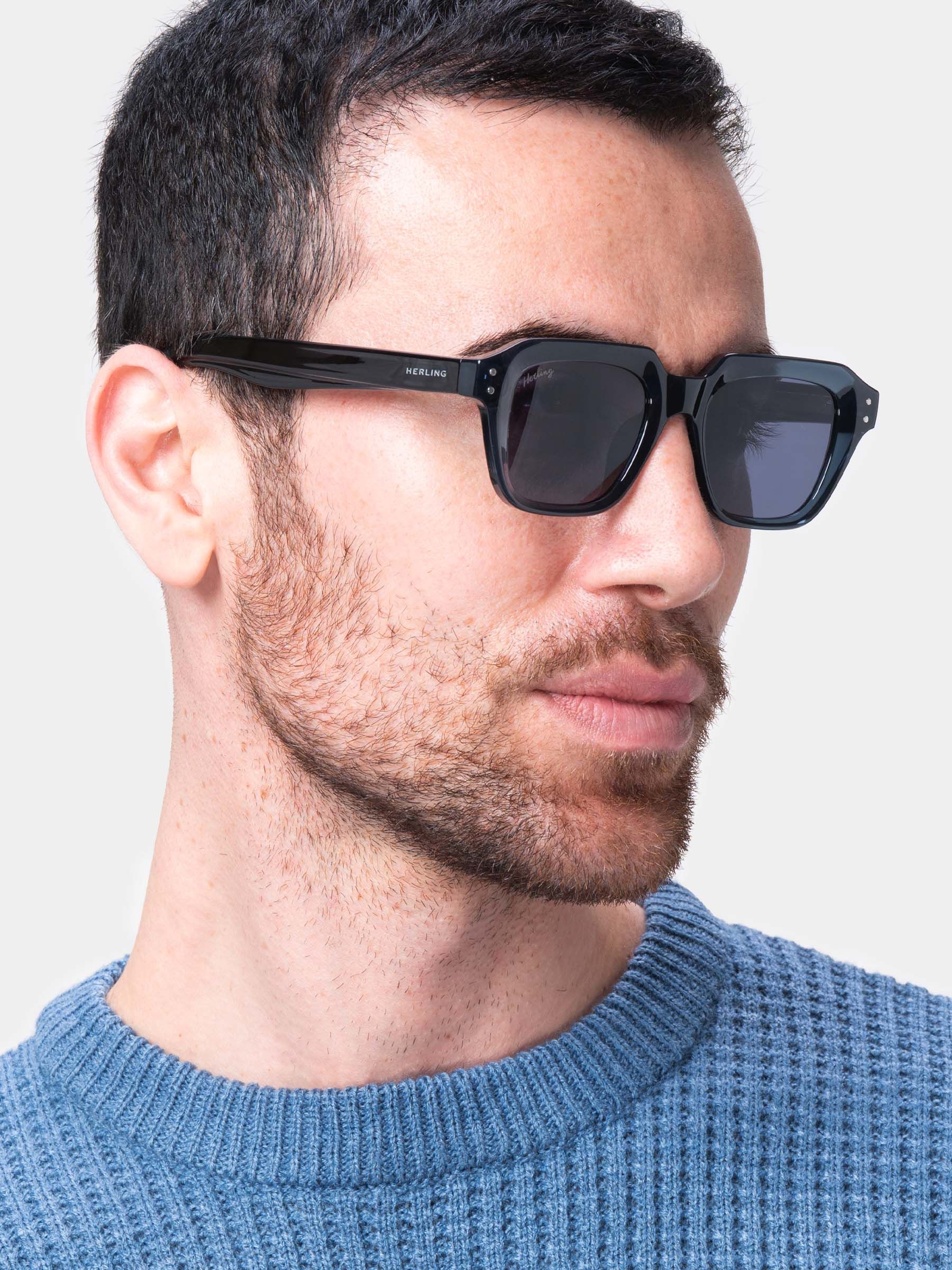 Uomo con occhiali da sole blu scuro. Logo Herling sulla stanghetta. Lenti quadrate. Maglione azzurro.